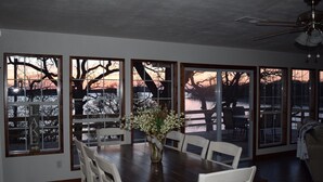 A dining area with a large table and chairs, a vase with flowers, and a view of a lake at sunset through large windows. - Allure 1900 sq waterfront home with country elegance spectacular views (Eastland)