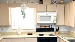 Una cocina con armarios de madera clara, un microondas blanco, una estufa y una ventana con una planta colgante.