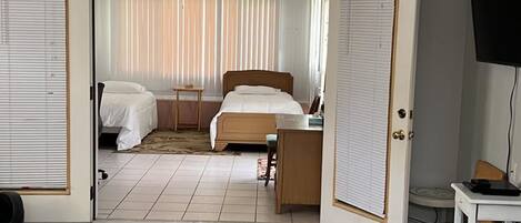 A room with a bed, a desk, a television, and a large window with blinds.