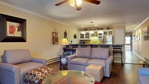 Una sala de estar con sofá, sillones y una mesa de centro de vidrio. Se observa una cocina al fondo, con armarios y una zona de comedor.