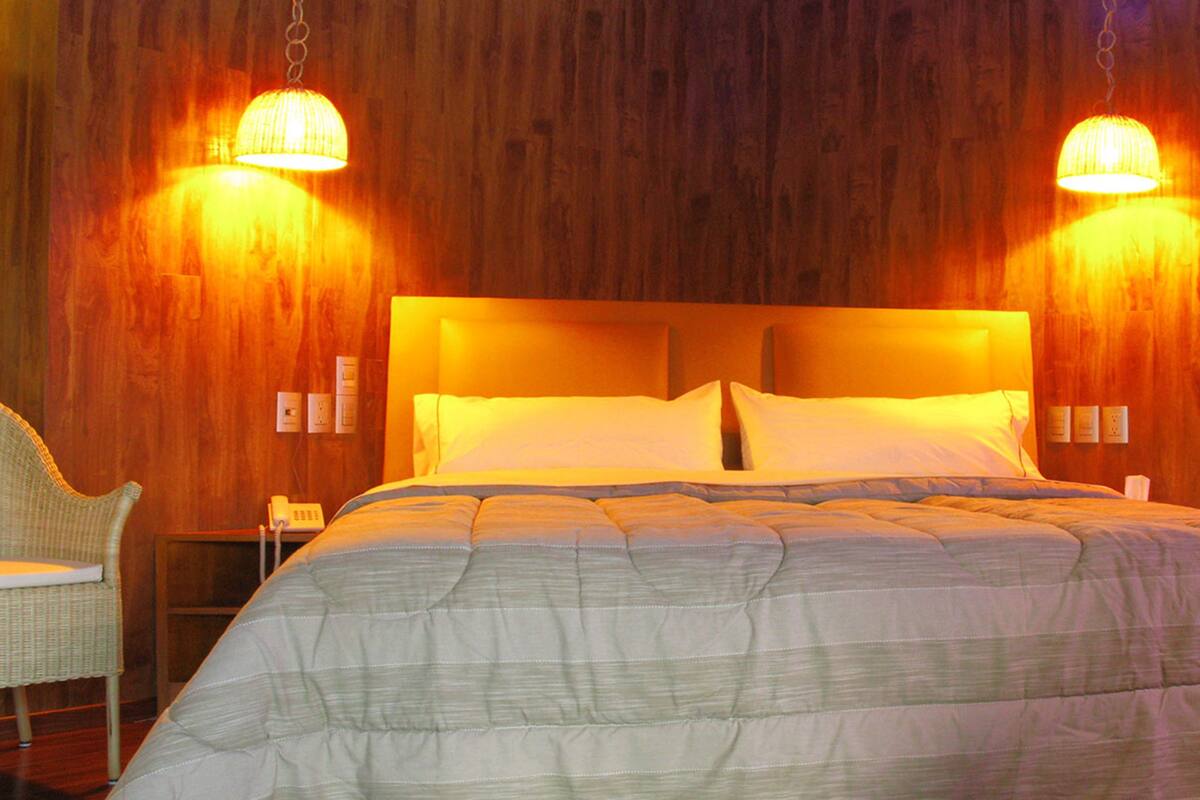 A hotel room with a large bed, two wall-mounted lamps, a wicker chair, and a nightstand with a phone.