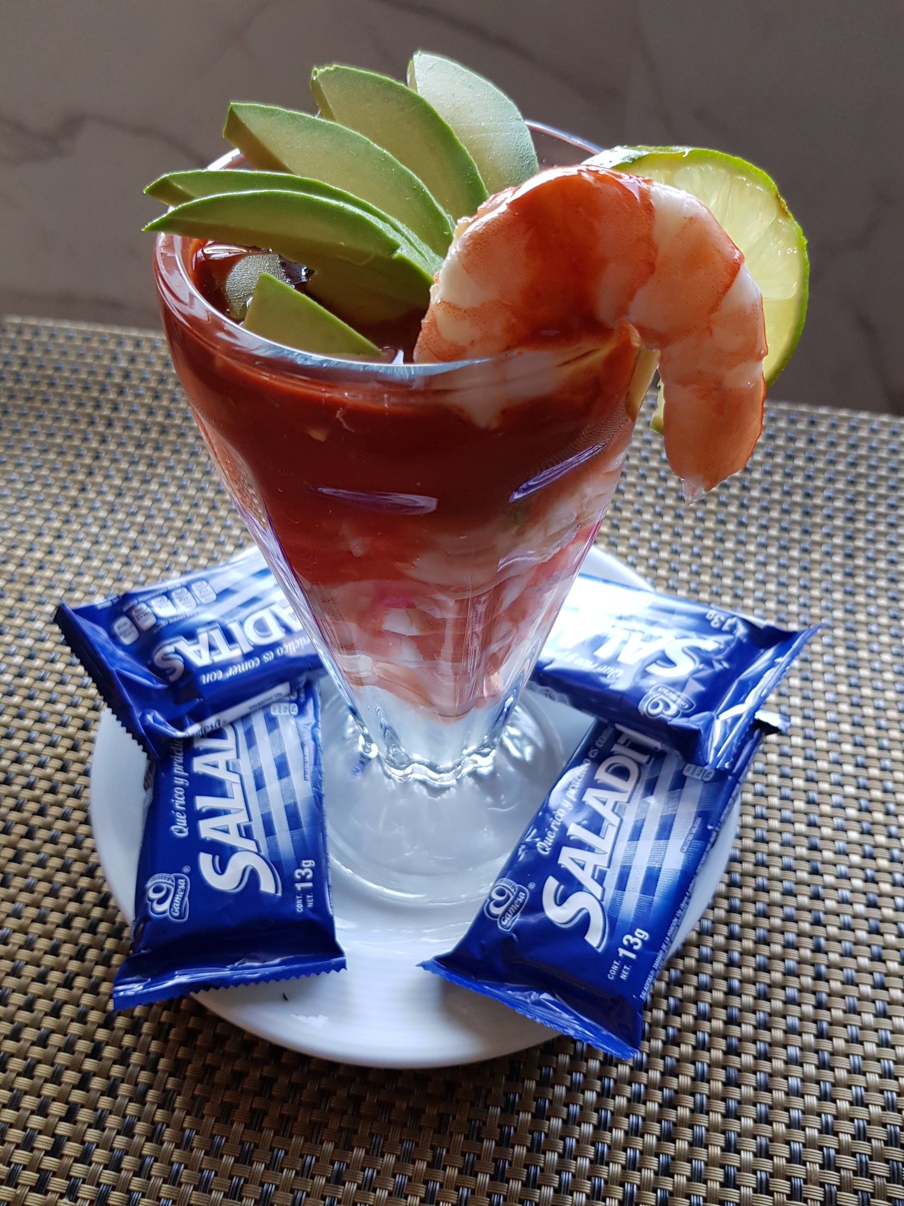 A cocktail glass with a shrimp and avocado garnish, accompanied by two packets of SALADI snacks.