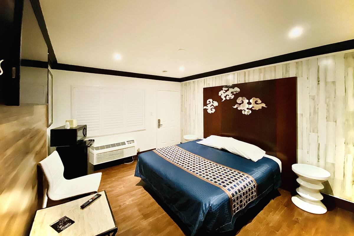 A modern hotel room with a large bed, a desk, a chair, a bedside table, and a decorative wall panel.