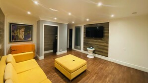 A modern living room with a yellow sofa, a small ottoman, a painting on the wall, and a television mounted on the wall. - Casa Blanca Inn & Suites (Whittier)