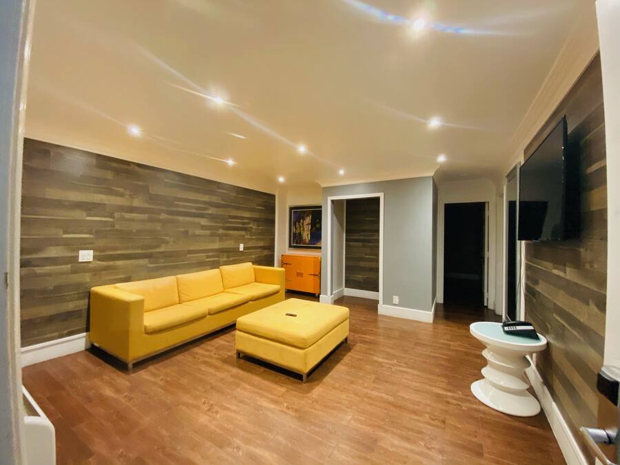 Una sala de estar moderna con un sofá seccional amarillo, un reposapiés blanco, piso de madera y un televisor de pantalla plana montado en la pared.