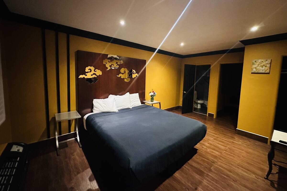 A modern hotel room with a large bed, a nightstand, a small table with a vase, and a bathroom door.