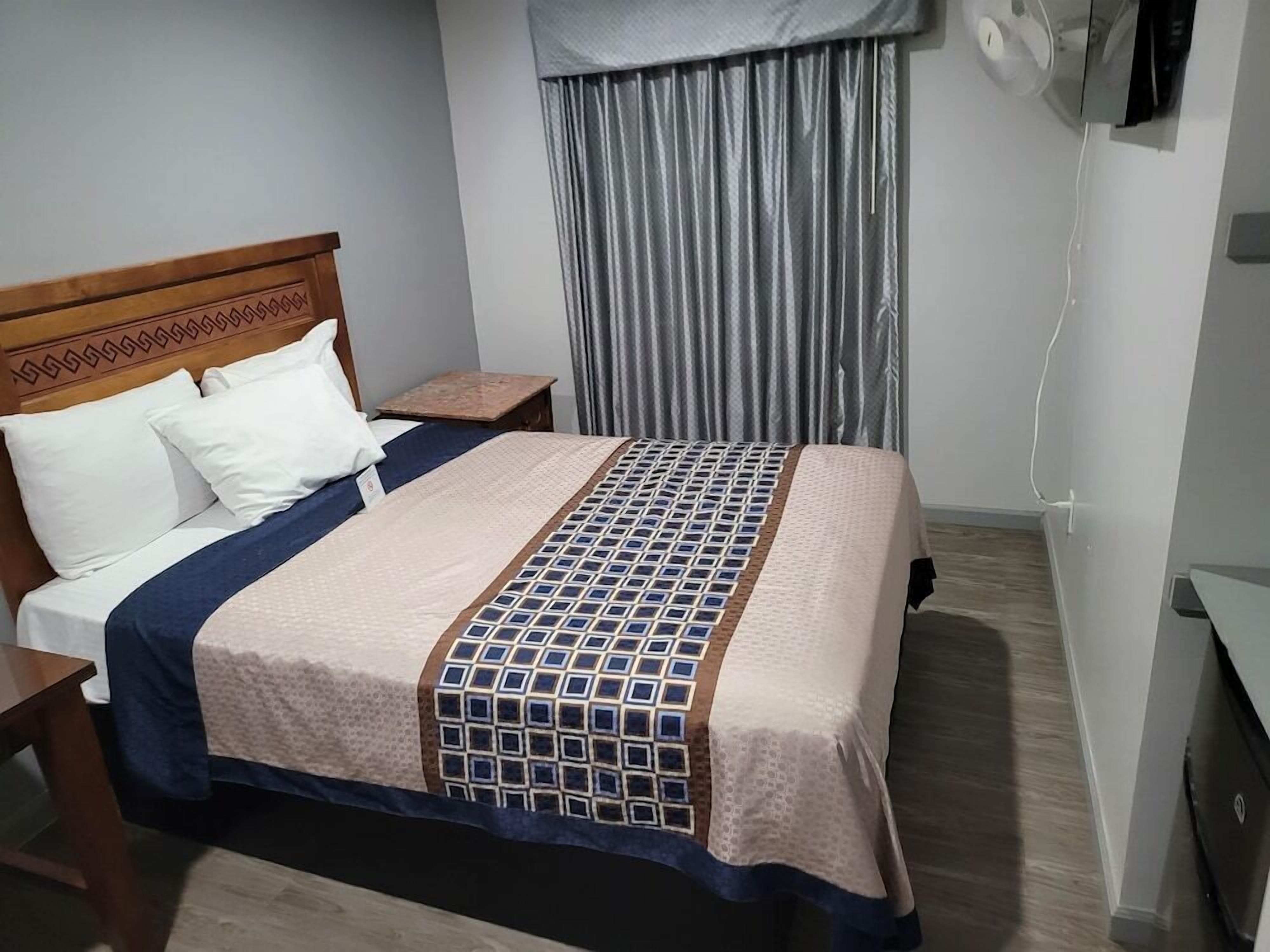 Une chambre d’hôtel avec un lit, une tête de lit en bois, une table de chevet, une télévision et une fenêtre avec des rideaux.
