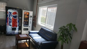 A room with a blue leather sofa, a wooden coffee table, a potted plant, a large Pepsi vending machine, and a window with a view of greenery.