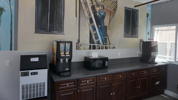 A kitchen with a coffee maker, microwave, and a refrigerator. There is a painting of two people on a ladder on the wall.
