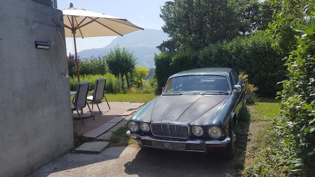 A vintage car parked in a garden with a concrete wall, outdoor chairs, and a table under a large umbrella. - Gästehaus Udligenswil (Udligenswil)