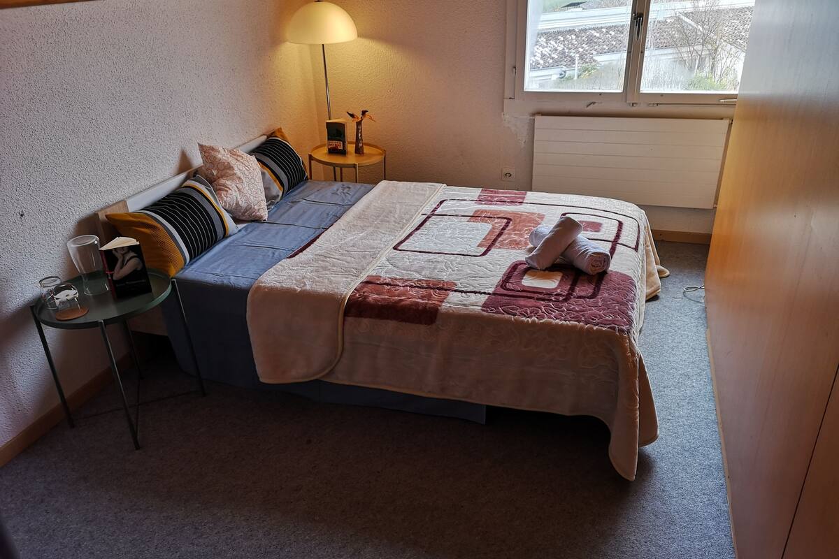 A bedroom with a bed, bedside table, lamp, and a view of the outside.