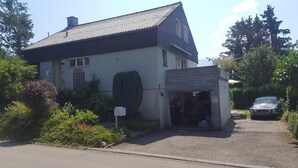 A two-story house with a garage entrance, surrounded by greenery and a parked car. - Gästehaus Udligenswil (Udligenswil)