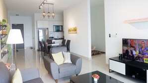 A modern living room with a flat-screen TV, grey sofas, and a dining area with a kitchen in the background. - FlexiAsia City Garden. Pinnacle Tower (Johor Bahru)