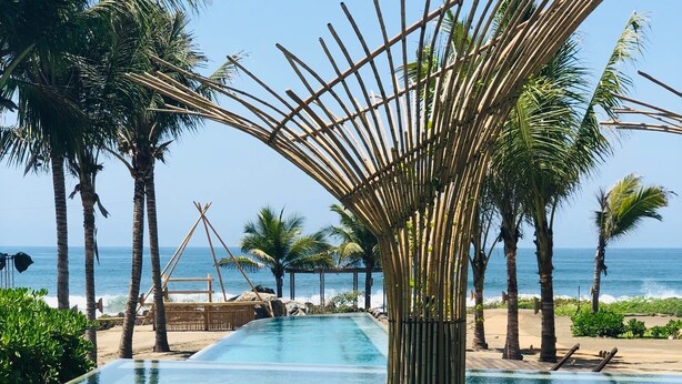 Une piscine aux eaux cristallines, agrémentée d’une structure en bambou au centre, entourée de palmiers et offrant une vue sur l’océan.