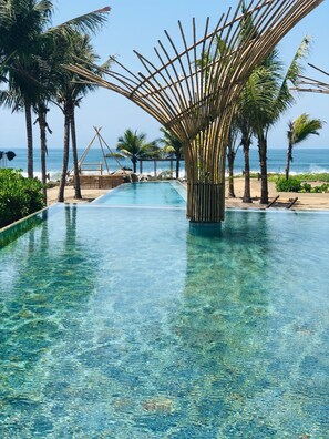 A clear pool with a bamboo structure in the center, surrounded by palm trees and a view of the ocean.