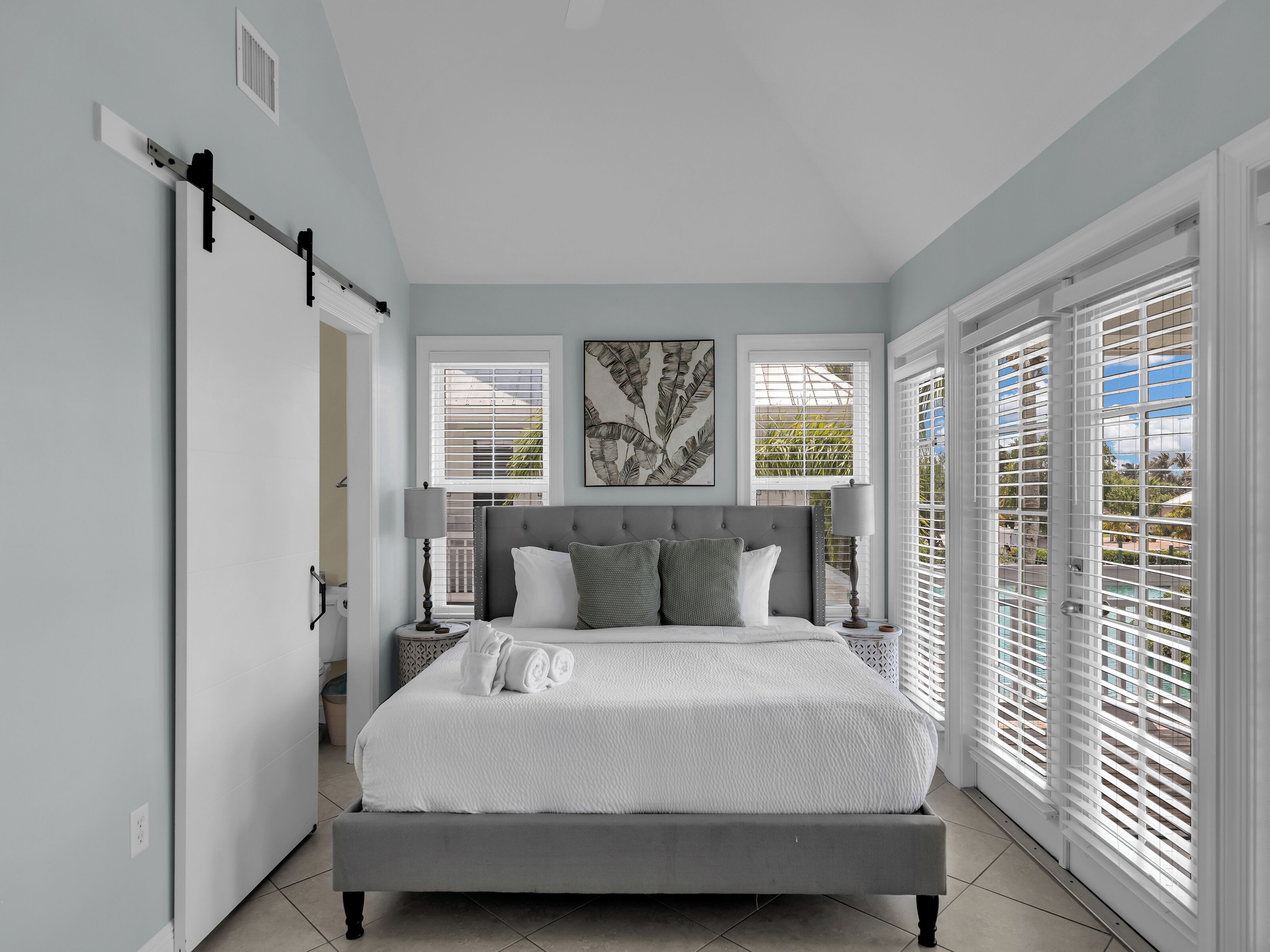 A bedroom with a large bed, a window with blinds, a bedside table with a lamp, and a wall-mounted shelf.
