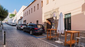 Een straat met kinderafvalsteen bestraat, met geparkeerde auto's en een rij gebouwen, waaronder een café met terras.