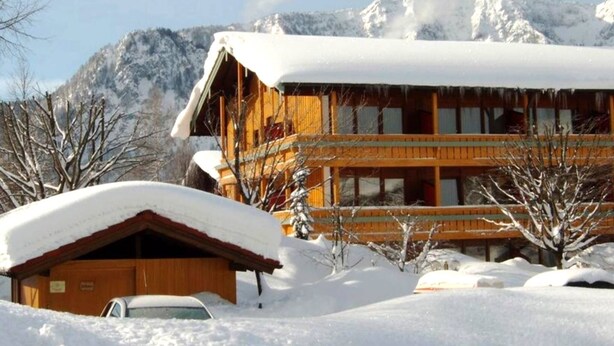 A wooden lodge with a snow-covered roof, surrounded by snow and mountains. - Ferienwohnung für 4 Personen, 2 Schlafzimmer mit Balkon, 60 m² (Inzell)