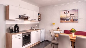 A modern kitchen with white cabinets, a built-in oven, and a coffee maker. There is a dining area with a table, chairs, and a red bench. A painting of a harbor scene hangs on the wall. - Ferienwohnung für 4 Personen, 2 Schlafzimmer mit Balkon, 60 m² (Inzell)