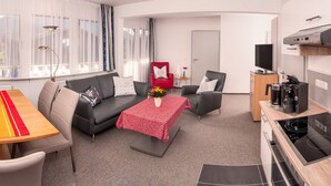 A modern living room with a sofa, a dining table, and a kitchen area with a stove. - Ferienwohnung für 4 Personen, 2 Schlafzimmer mit Balkon, 60 m² (Inzell)