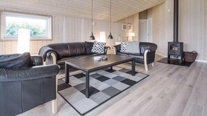 A modern living room with a black leather sofa, a coffee table, and a black and white checkered rug. There is a black wood-burning stove and a window with a view of greenery.