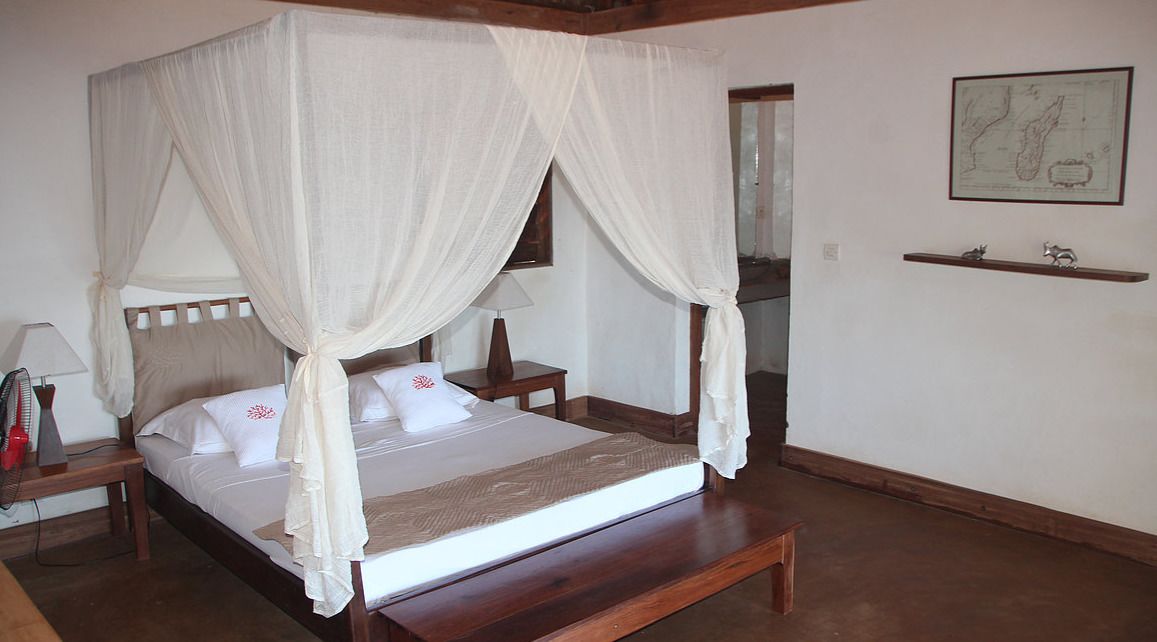 A four-poster bed with white linens and a canopy, a bedside table with a lamp, and a small shelf with decorative items.