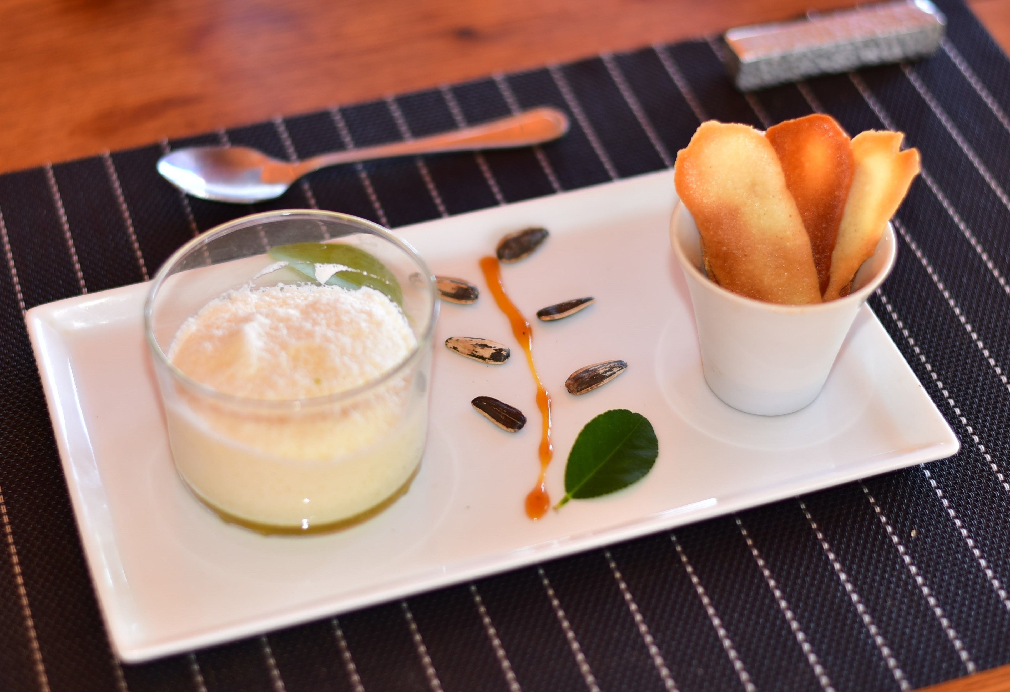 Un plato de postre con un vaso de natillas, una taza pequeña de pan y elementos decorativos como semillas y una hoja.