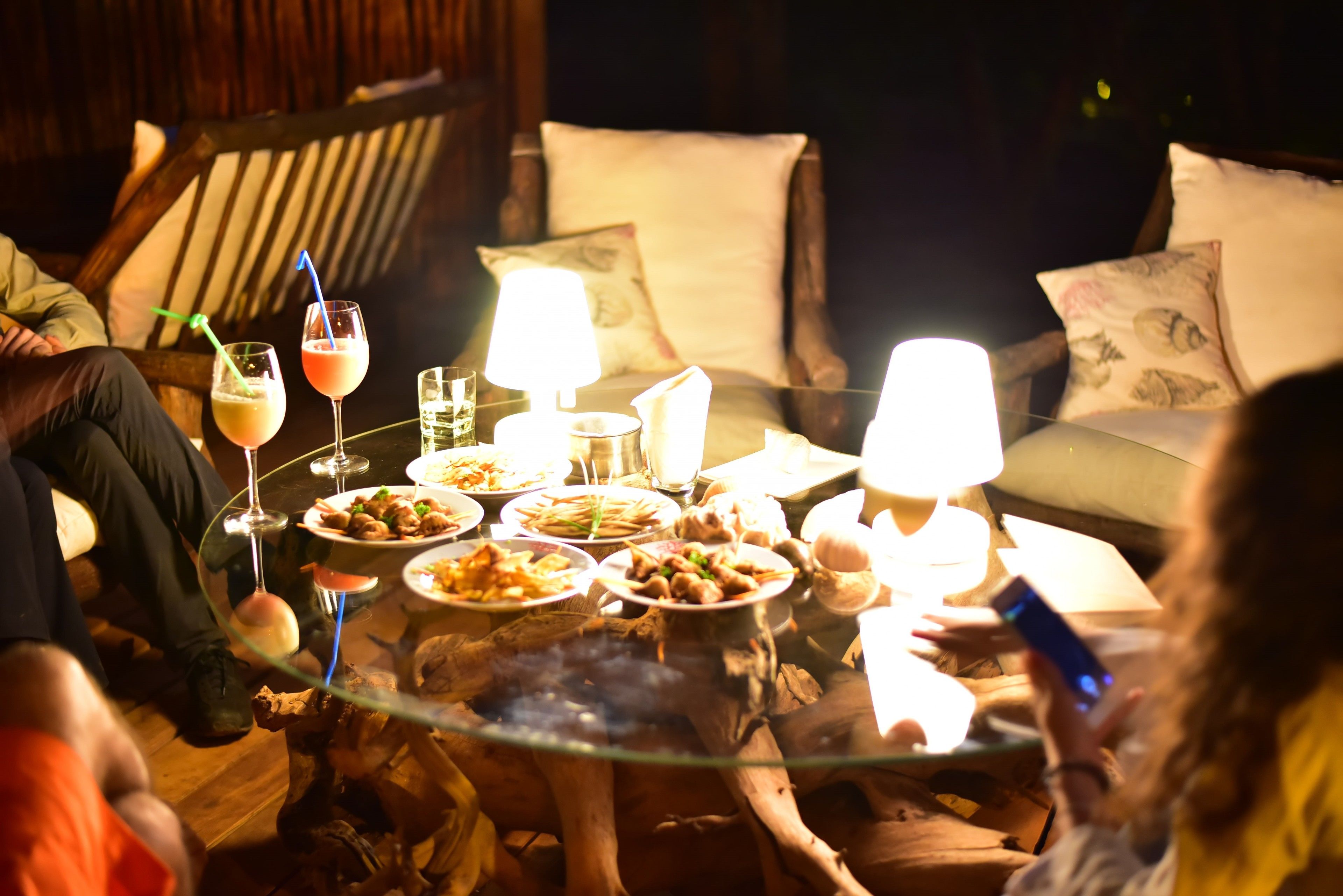 A round glass table with multiple plates of food and drinks, surrounded by wooden chairs with cushions.