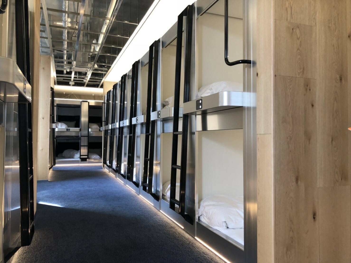 A corridor with bunk beds, a wooden wall, and a ceiling with exposed pipes.