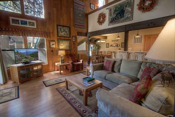 A spacious living room with a large sofa, a television, and a fireplace. There are several framed pictures on the walls and a wooden cabinet.