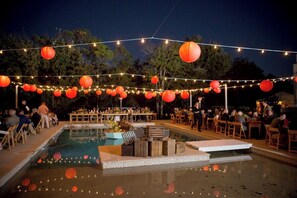 Un espace extérieur avec une piscine, agrémenté de guirlandes lumineuses et de lanternes, et entouré de mobilier et de tables en bois.