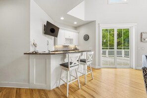 A kitchen with white cabinetry, a marble countertop, and two bar stools. There is a large window with a view of greenery outside.
