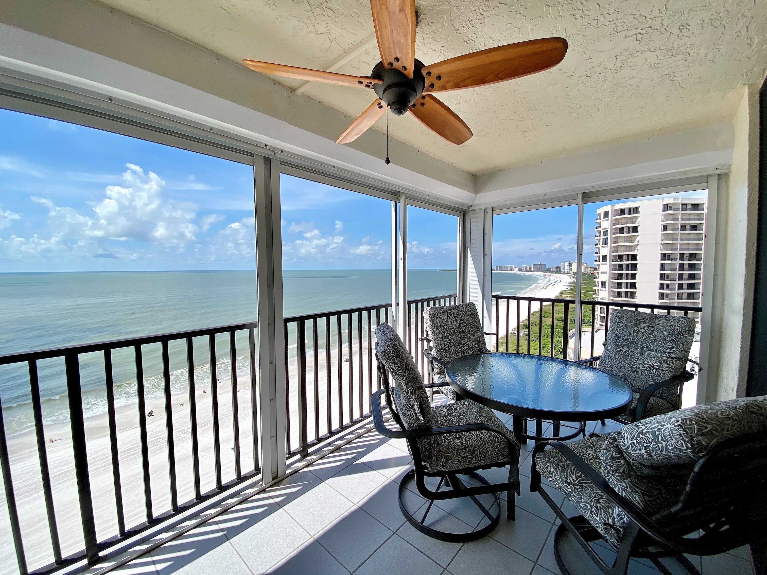 A balcony with a table and chairs, a ceiling fan, and a view of the ocean and beach.