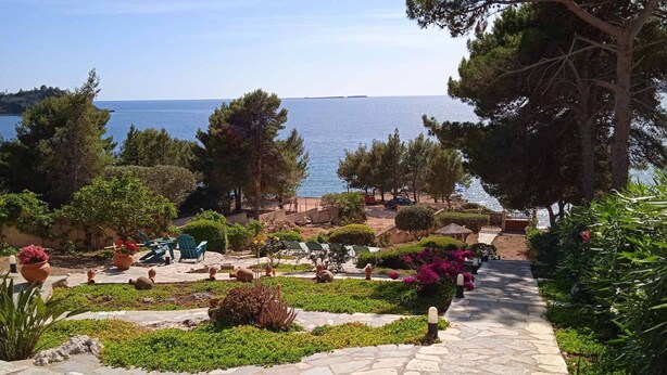 Ein gepflasterter Weg führt zu einer Sitzgruppe mit Stühlen und Tischen, umgeben von üppiger Vegetation und Blick aufs Meer.