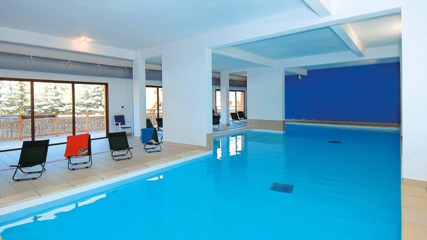 En indendørs swimmingpool med klart, blåt vand, omgivet af hvide vægge og store vinduer.