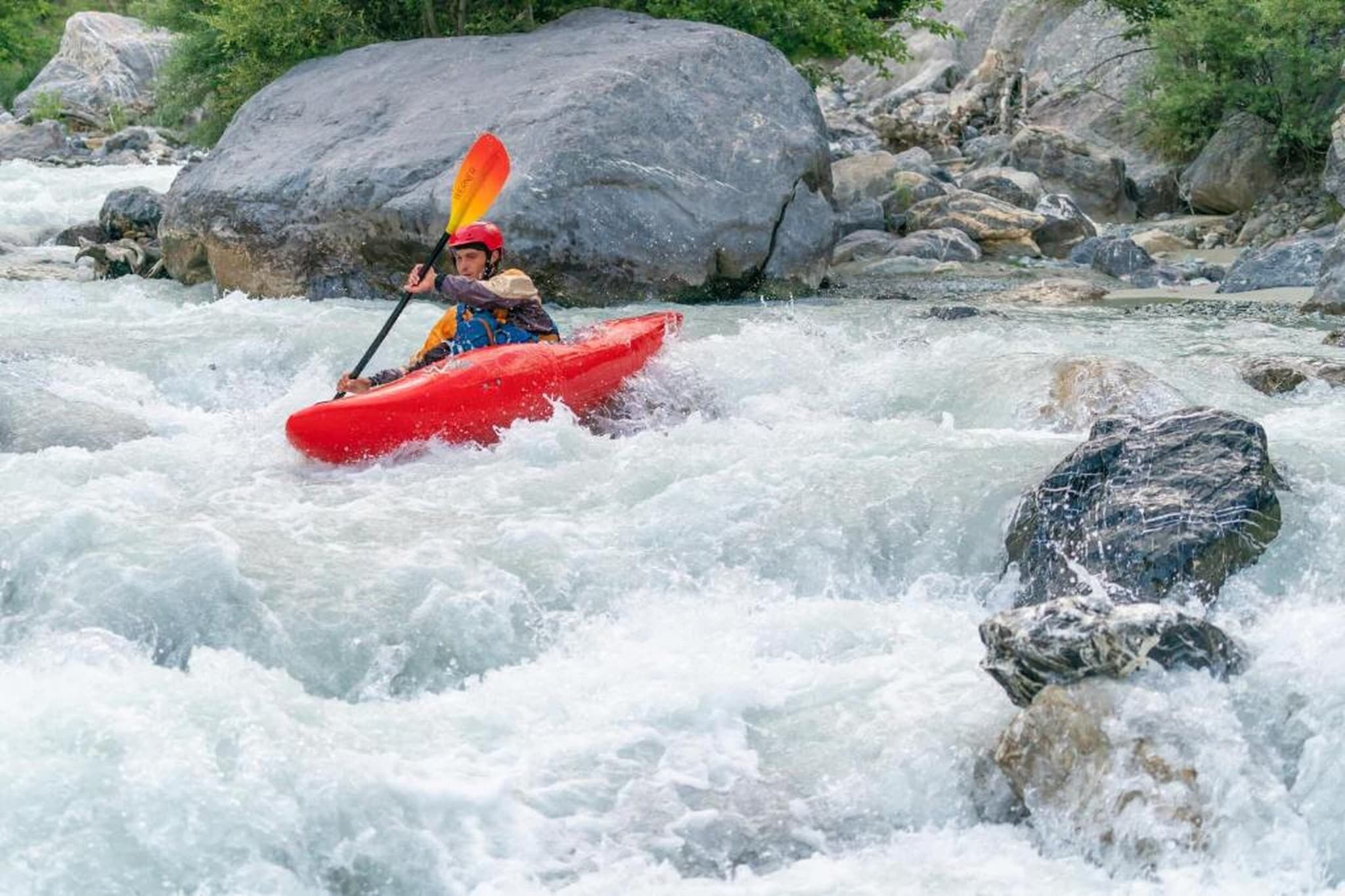 A person kayaking on a river with rapids, surrounded by rocks and greenery.
