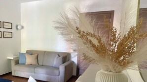 Uma sala de estar moderna com um sofá cinzento, um vaso branco com plantas secas e quadros nas paredes.