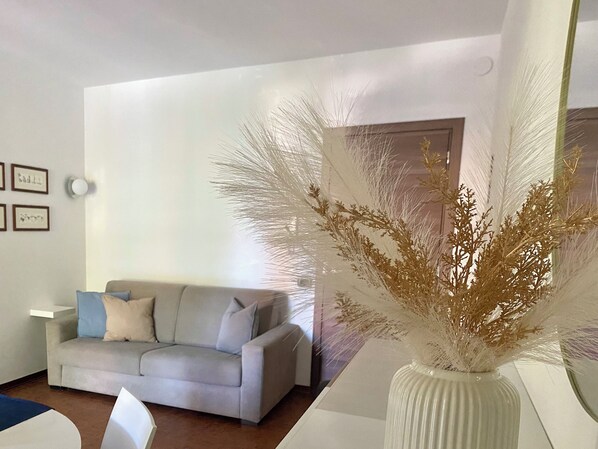 A modern living room with a grey sofa, a white vase with dried plants, and framed pictures on the wall.