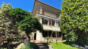 Um edifício de dois andares com varanda, rodeado por jardins bem cuidados e um caminho pavimentado.