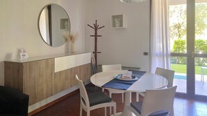 Uma sala de jantar com uma mesa redonda, cadeiras brancas, um aparador de madeira, um porta-roupas, um lustre e uma porta de vidro deslizante que dá para uma área externa.