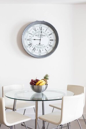 A round glass table with four white chairs, a bowl of fruit, and a large wall clock. - Rooms & Suites Loft 2H - Arrecife, Lanzarote (Arrecife)