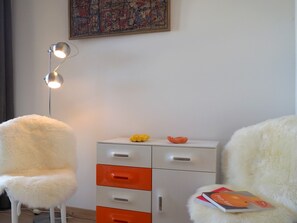 Una habitación con una silla blanca, un armario blanco con cajones naranjas, una lámpara de pie y un cuadro en la pared.