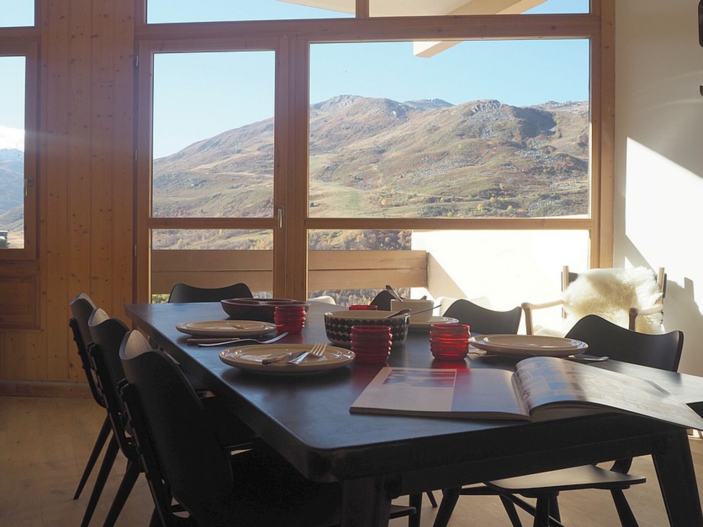 Un coin repas avec une table dressée pour quatre personnes, de grandes fenêtres donnant sur les montagnes et un objet blanc et moelleux sur la droite.