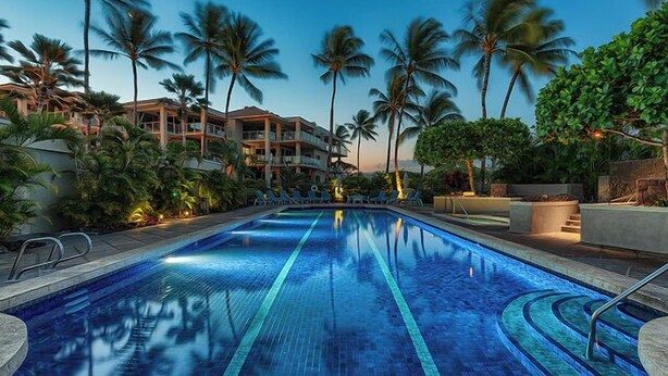 A swimming pool with clear blue tiles, surrounded by palm trees and a building with multiple balconies.