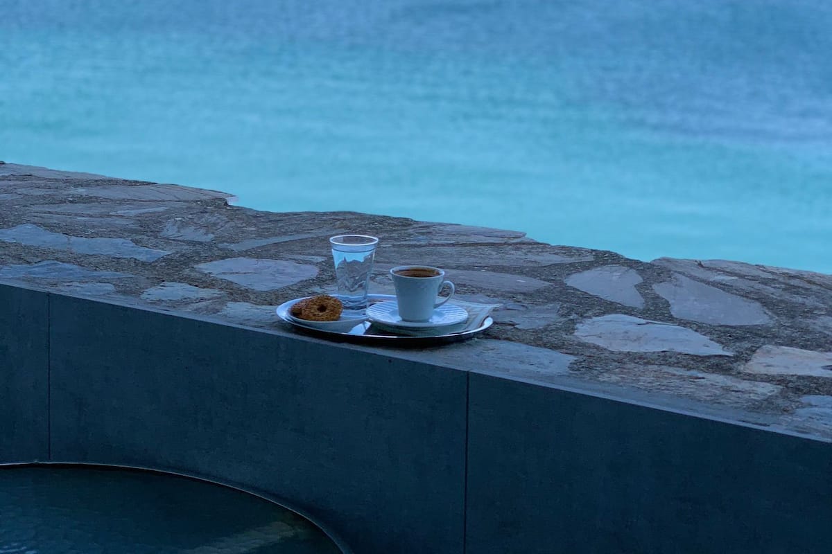 Una mesa con una taza y platillo, un vaso y un pastel sobre un saliente de piedra con vista a un cuerpo de agua.
