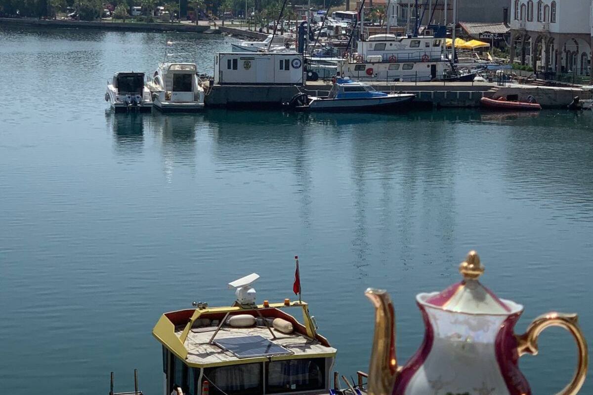 Un puerto deportivo con barcos amarrados, una mesa con juego de té, y una vista panorámica de colinas al fondo.