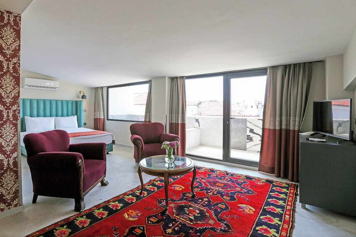 Una habitación de hotel con una cama, dos sillones, una mesita con un jarrón, televisión y un amplio ventanal con cortinas.