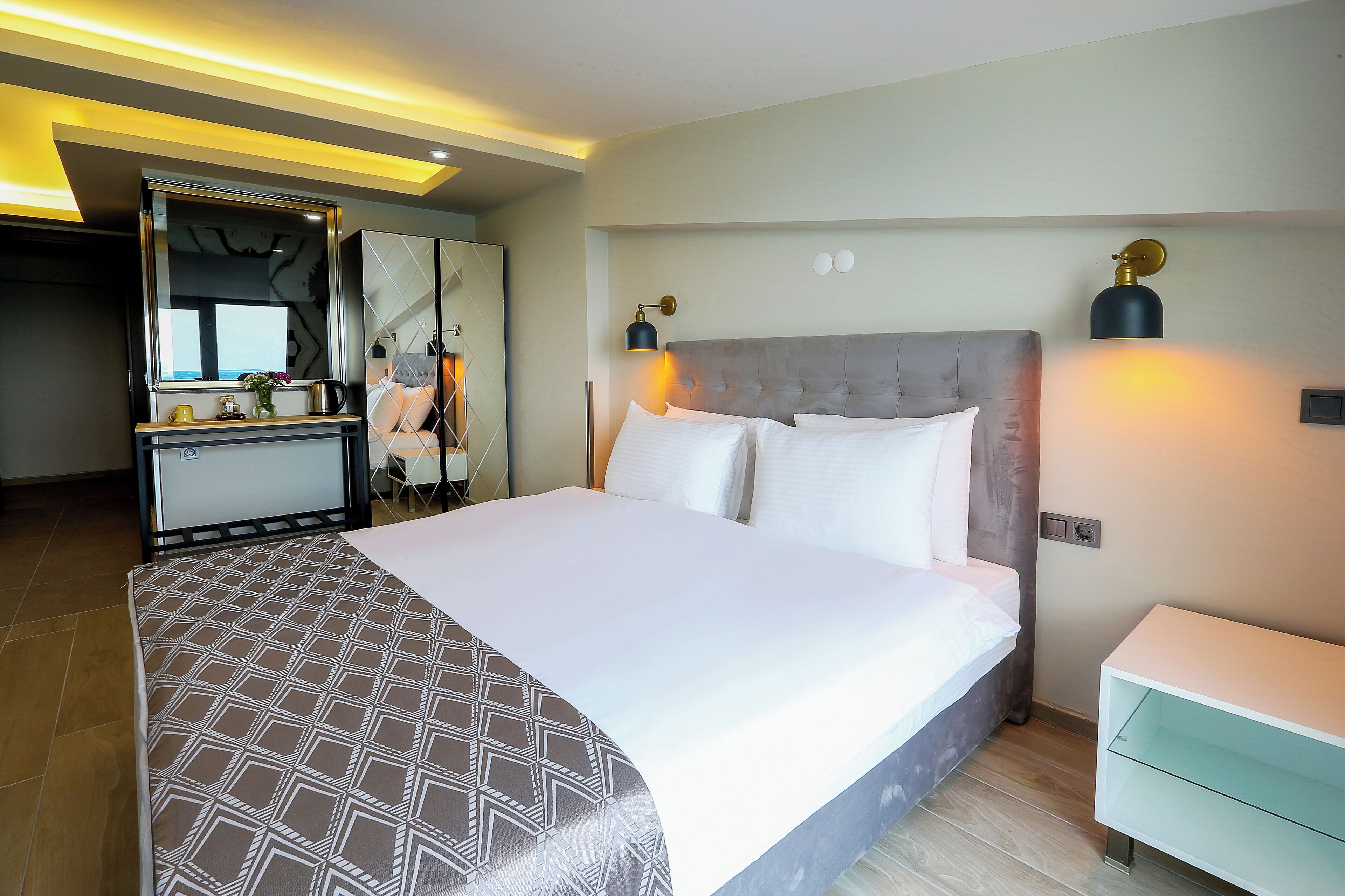 Una habitación de hotel moderna con una cama grande, mesitas de noche, un tocador con espejo e iluminación empotrada.