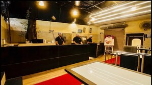 Interior de restaurante con una larga barra, tres cocineros al fondo y una alfombra roja que lleva al área de mesas.