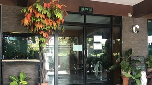 A hotel entrance with a digital clock displaying 8:59:00, a glass door, and potted plants. - Lert Sri Hotel (Suphan Buri)
